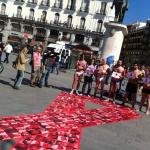 En la Puerta del Sol de Madrid cinco hombres en calzoncillos y una mujer vestida sostienen un cartel con el lazo rojo del sida, en el suelo hay un lazo grande rojo con rostros de personas 