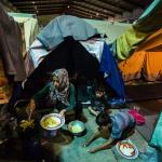 Una mujer cocina junto a dos niños frente a una tienda de campaña en un campo de refugiados griego.